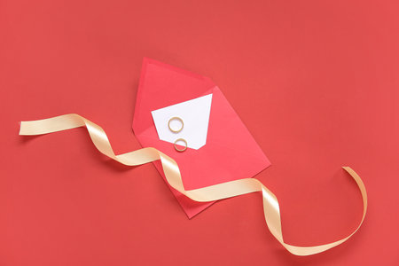 Envelope with greeting card, rings and golden ribbon on red backgroundの写真素材