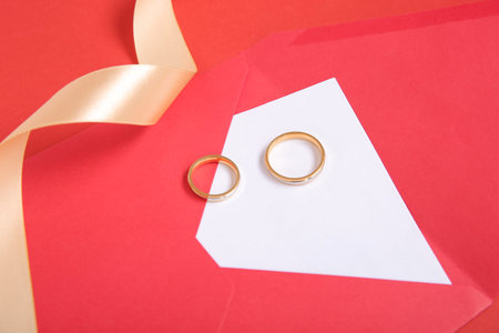 Envelope with greeting card, rings and golden ribbon on red backgroundの写真素材
