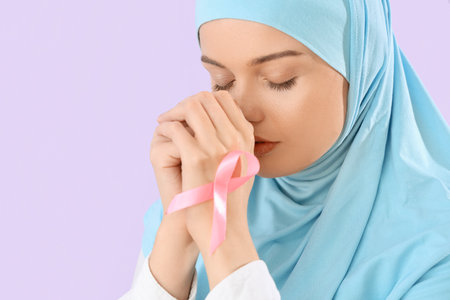 Young Muslim woman with pink ribbon praying on lilac background, closeup. Breast cancer awareness conceptの写真素材