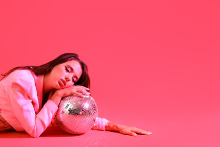 Fashionable beautiful young woman with disco ball lying on red backgroundの写真素材