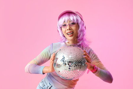 Happy fashionable young Asian woman with disco ball on pink background