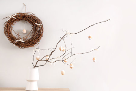 Vase with tree branches, Easter eggs and bows on table near light wall in room