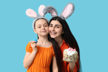 Happy smiling young woman with her daughter in bunny ears holding Easter cake and egg on blue backgroundの写真素材