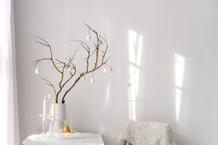 Vase with tree branches, Easter eggs and burning candles on table in light roomの写真素材