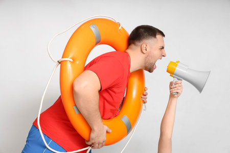 Young lifeguard with lifebuoy shouting into megaphone on white background
