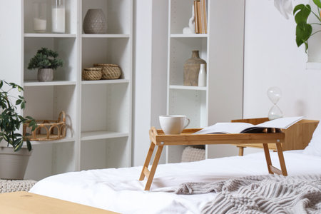 Wooden stand with coffee cup and magazine on bed in light roomの写真素材