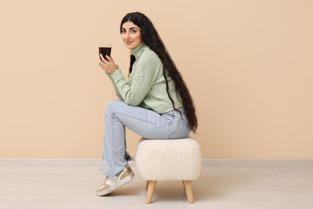 Beautiful young woman with glass cup of hot tea sitting on pouf near beige wallの写真素材