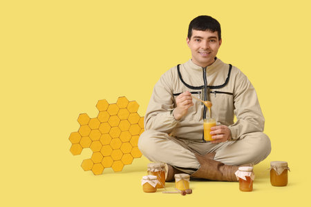 Male beekeeper with honey and paper honeycombs sitting on yellow backgroundの写真素材