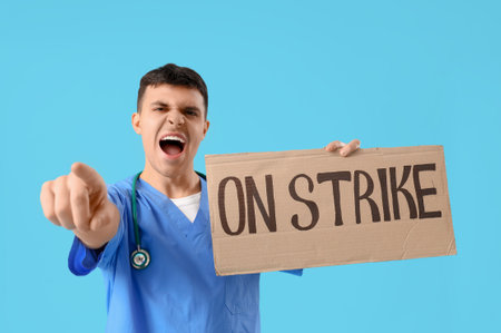 Protesting male doctor holding placard with text ON STRIKE and pointing at viewer against blue backgroundの写真素材