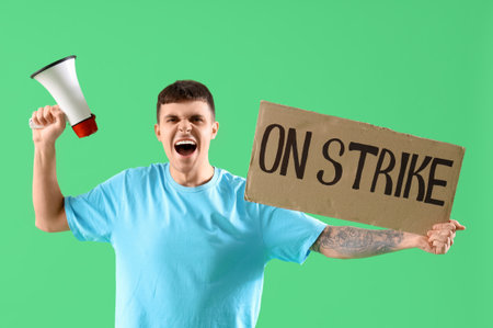 Protesting young man holding placard with text ON STRIKE and megaphone against green backgroundの写真素材