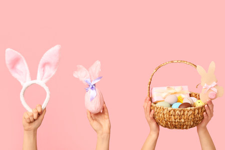 Female hands holding basket with Easter eggs, bunny ears and gift on pink backgroundの写真素材