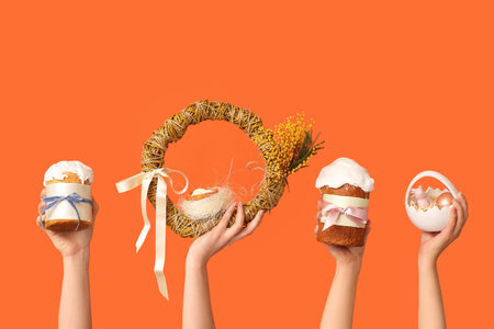 Female hands holding Easter cakes with wreath and basket on orange background