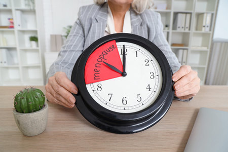 Mature businesswoman holding clock with the word MENOPAUSE in office, closeup
