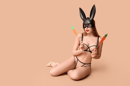 Sexy young woman in bunny mask with paper carrots sitting on beige background. Easter celebrationの写真素材