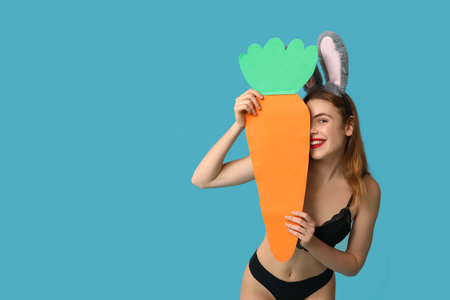 Sexy smiling young woman in bunny ears with paper carrot on blue background. Easter celebrationの写真素材