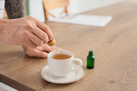 Young man dropping CBD oil into tea cup in kitchen, closeup