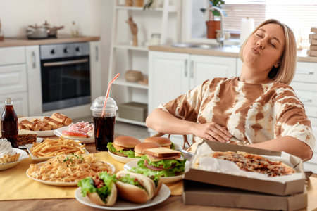 Beautiful woman at table full of unhealthy food in kitchen. Overeating conceptの写真素材