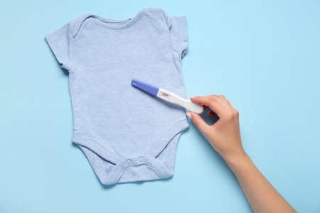 Female hand with positive pregnancy test and baby bodysuit on color background
