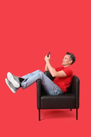 Handsome man using mobile phone in black armchair on red backgroundの写真素材