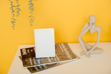 Blank frame, magazines and decorative figurine on white table near orange wall, closeupの写真素材