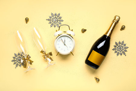 Alarm clock with Christmas balls, glasses and bottle of champagne on yellow backgroundの写真素材