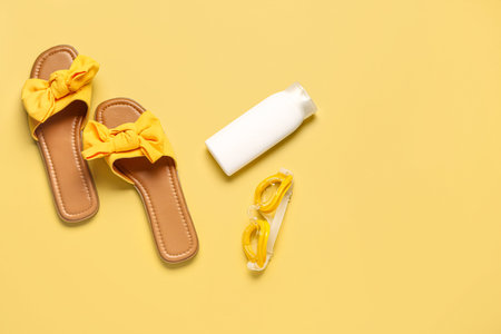 Female stylish flip flops with sunscreen cream and goggles on yellow background