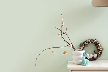 Vase with tree branches, Easter eggs and wreath on shelf near light wall