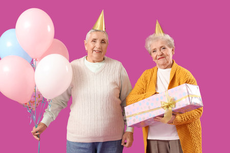 Senior female friends in birthday hats with balloons and gift on purple backgroundの写真素材