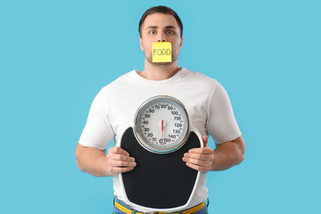 Young man with taped mouth and scales on blue background. Weight loss conceptの写真素材