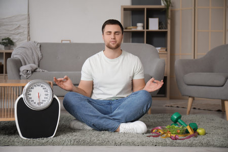 Young man with scales, dumbbells and food meditating at home. Weight loss conceptの写真素材
