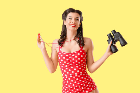 Beautiful pin-up lifeguard with whistle and binoculars on yellow backgroundの写真素材
