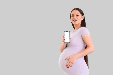 Young pregnant woman with medical patch and mobile phone after vaccination on light background
