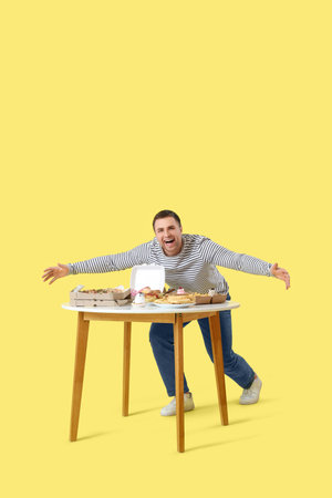 Young man with unhealthy food on table against yellow background. Overeating conceptの写真素材