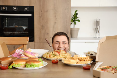 Young man behind table with unhealthy food in kitchen. Overeating conceptの写真素材