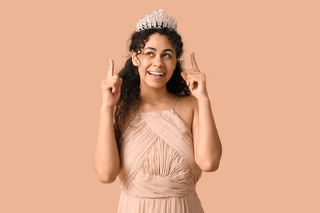 Beautiful young African-American woman in stylish beige prom dress pointing at tiara on brown backgroundの写真素材