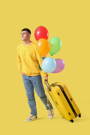 Young man with suitcase and bunch of balloons on yellow backgroundの写真素材