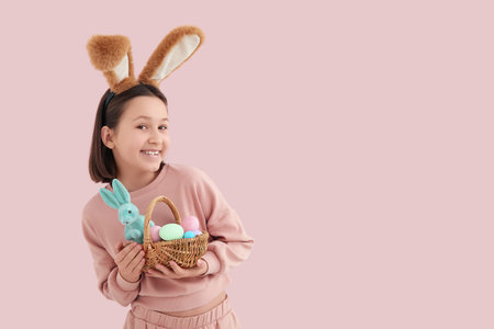 Happy little girl in bunny ears holding basket with Easter eggs and rabbit on pink background