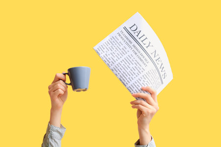 Female hands with newspaper and cup of coffee on yellow backgroundの写真素材