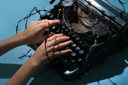 Female hands with vintage typewriter and barbed wire on blue background. Printing ban concept