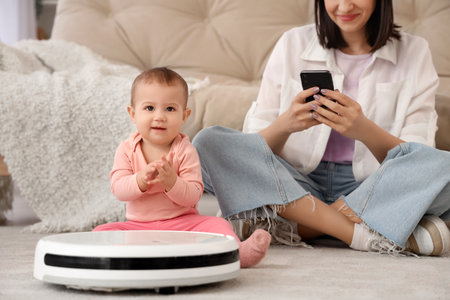 Cute little baby with robot vacuum cleaner and mother using mobile phone on carpet at homeの写真素材