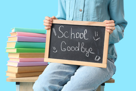 Female student sitting on desk and holding chalkboard with text SCHOOL GOODBYE on blue background. End of school conceptの写真素材