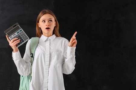 Surprised female student with calculator pointing at something on blackboard background. End of school conceptの写真素材
