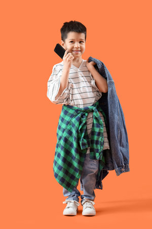 Cute little Asian boy in adult clothes talking by mobile phone on orange backgroundの写真素材