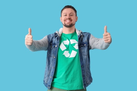 Happy young man in t-shirt with recycling logo showing thumbs-up on blue background. Ecology conceptの写真素材