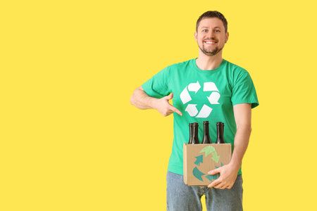 Happy young man in t-shirt with recycling logo pointing at cardboard box with glass bottles on yellow background. Ecology conceptの写真素材