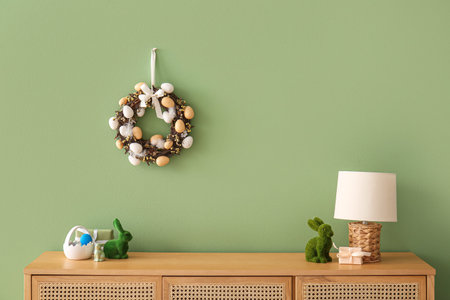 Beautiful Easter wreath decorated with eggs on green wall near chest of drawers with toy bunnies in...