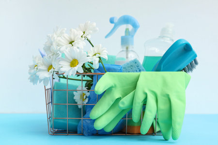 Basket with cleaning supplies, tools and daisies on blue table against white background