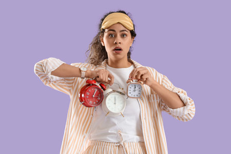 Oversept young African-American woman in pajamas with alarm clocks on lilac background
