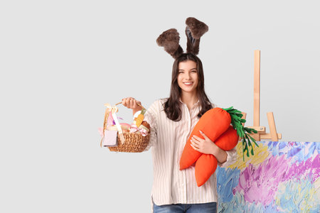 Happy female artist in bunny ears with Easter basket, toy carrots and easel on gray background