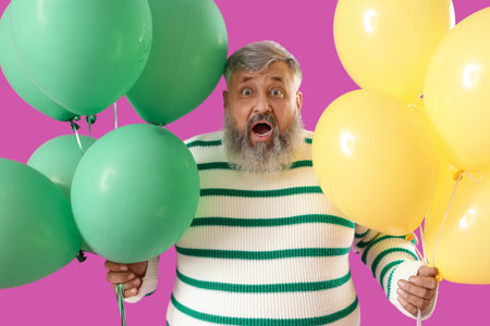 Surprised senior man with bunches of colorful balloons on pink backgroundの写真素材